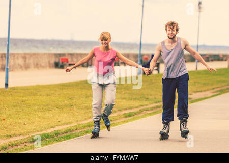 Aktive Menschen moderne Paar zusammen trainieren und Spaß haben. Entspannendes Hobby. Junge Menschen halten Hände beim Inline-Skating im Park. Stockfoto