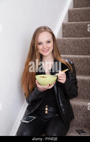 Eine langhaarige rothaarige Frau sitzt auf der Treppe ihres Hauses Getreide aus einer Schüssel essen. Stockfoto