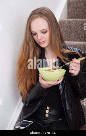 Eine langhaarige rothaarige Frau sitzt auf der Treppe ihres Hauses Getreide aus einer Schüssel essen, während ihr Handy betrachten. Stockfoto