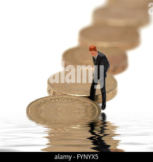 Ein Miniatur-Business-Mann in einem Anzug mit Blick auf einige Pfund-Münzen mit einem Digital reflektierten Wasserbecken. Stockfoto