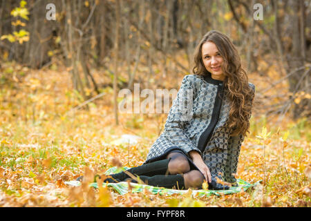 Junge schöne Mädchen in einem gelben Mantel sitzt auf Laub im Wald Stockfoto