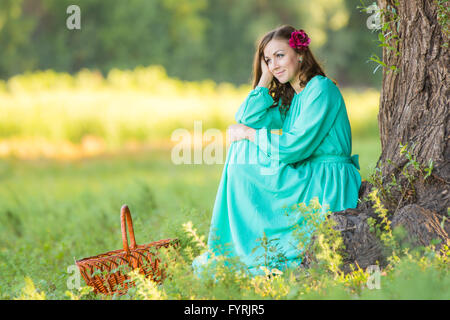 Eine Mädchen in einem langen Kleid und setzte sich bei dem Gedanken an einen alten Baum im Wald Stockfoto