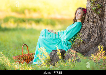 Eine Mädchen in einem langen Kleid setzte sich an den alten Baum im Wald Stockfoto