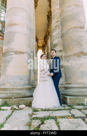 Hübscher Bräutigam und seine schöne Braut im weißen Kleid stehen Hand in Hand zwischen zwei Spalten bei Sonnenuntergang Stockfoto