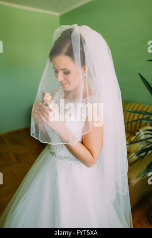 Schöne Braut in Spitze Hochzeitskleid und Schleier betrachten das Knopfloch der Rosen im Haus Stockfoto