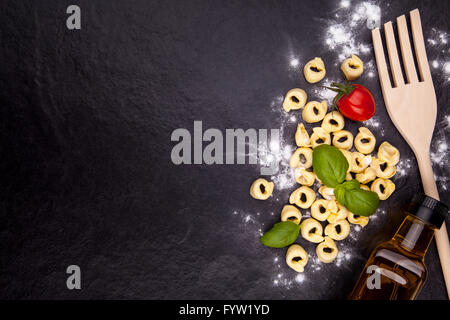 Tortellini, Tomaten, Basilikum, Olivenölflasche und einem Holzlöffel auf einem schwarzen Steinplatte. Draufsicht mit Exemplar Stockfoto