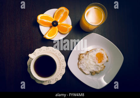 Frühstück auf der dunklen Oberfläche des Holzes inklusive Kaffee, Brot, Orangensaft, Spiegelei und Scheiben Orangen auf kleinen weißen Teller gelegt Stockfoto