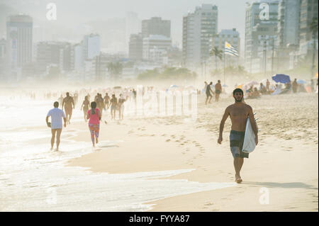 RIO DE JANEIRO - 18. Januar 2014: Surfer Spaziergänge am Strand von Ipanema Strand gegen einen nebligen Sonnenuntergang Blick auf die Skyline der Stadt Stockfoto