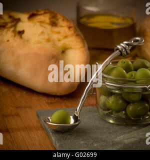Glas mit grünen Oliven mit einzelnen Olive auf Löffel und Olivenöl mit Brot im Hintergrund. Stockfoto