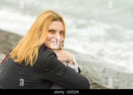 Porträt des Sitzens auf dem Strand lächelnde junge Frau in der kalten Jahreszeit Stockfoto