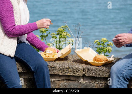 Zwei Personen Urlauber sitzen auf einem Meer wand Essen takeaway Fish und Chips aus Polystyrol Behälter in der Küstenstadt New Quay Wales UK Stockfoto