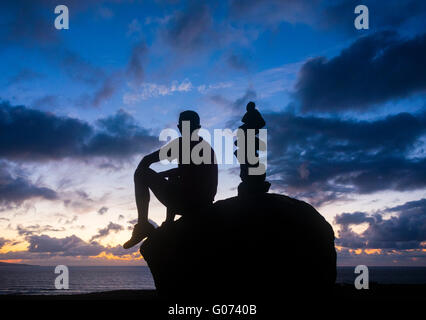 Mann mit Blick auf das Meer bei Sonnenuntergang sitzen auf Felsen in der Nähe von Steinen im Gleichgewicht zu halten. Stockfoto