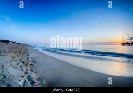 Die aufgehende Sonne lugt durch die Wolken und spiegelt sich in Wellen von dem Nags Head Fishing Pier auf den outer Banks von North Carolina Stockfoto