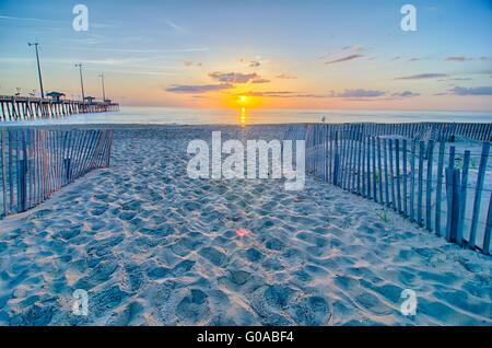 Die aufgehende Sonne lugt durch die Wolken und spiegelt sich in Wellen von dem Nags Head Fishing Pier auf den outer Banks von North Carolina Stockfoto