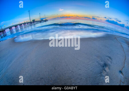 Die aufgehende Sonne lugt durch die Wolken und spiegelt sich in Wellen von dem Nags Head Fishing Pier auf den outer Banks von North Carolina Stockfoto