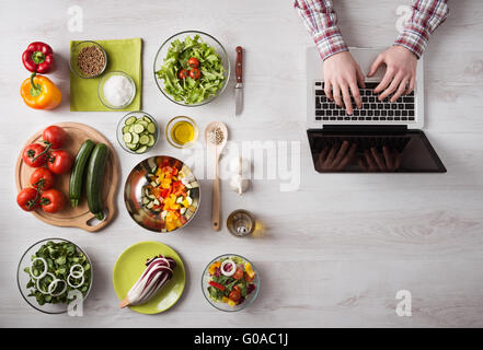 Mann in der Küche auf der Suche nach Rezepten auf seinem Laptop mit Lebensmittelzutaten und frisches Gemüse auf der linken, oberen Ansicht Stockfoto