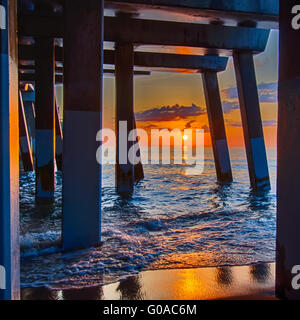 Die aufgehende Sonne lugt durch die Wolken und spiegelt sich in Wellen von dem Nags Head Fishing Pier auf den outer Banks von North Carolina Stockfoto