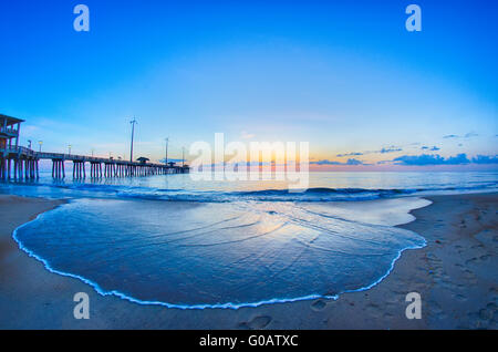 Die aufgehende Sonne lugt durch die Wolken und spiegelt sich in Wellen von dem Nags Head Fishing Pier auf den outer Banks von North Carolina Stockfoto