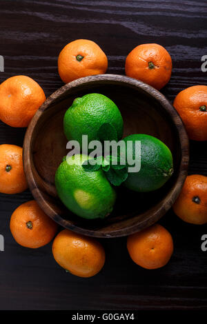 Limetten in Holzteller mit Mandarinen und Minze Blätter auf dunklem Hintergrund Nahaufnahme Makro Stockfoto