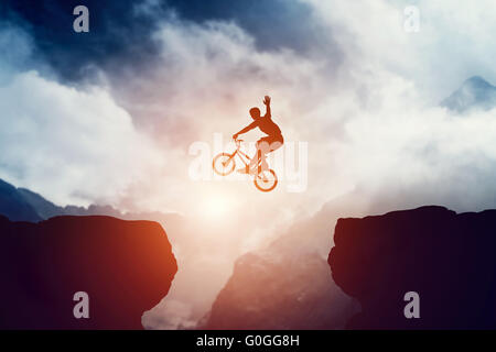 Mann springt auf bmx-Rad über Abgrund in Berge bei Sonnenuntergang. Erhöhung der Hand Hallo Geste zeigt. Extreme sport Stockfoto