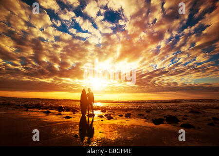 Surfer stehend mit seinem Surfbrett am Strand bei Sonnenuntergang über dem Ozean Stockfoto