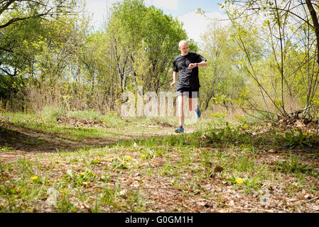 Ein senior Mann, getragen in schwarz ist in den Wald laufen und sieht zur Zeit auf seine Stoppuhr, während ein warmer Frühlingstag Stockfoto