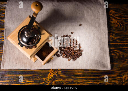 Top-down Ansicht auf quadratisch geformte hölzerne Kaffeemühle, Gelände und kleine Haufen von Bohnen auf Tischdecke Stockfoto