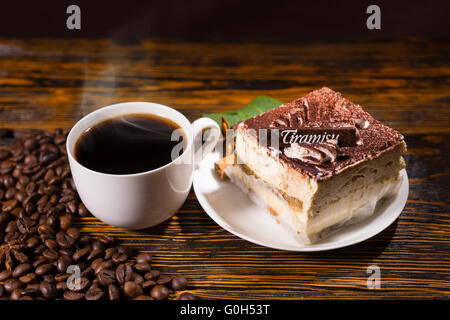 Köstliche quadratische Kuchen Stück in Platte neben volle Tasse Kaffee umgeben von dunklen Bohnen über gebeiztem Holz Hintergrund Stockfoto