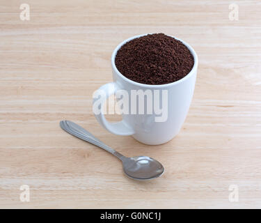Eine Tasse gefüllt mit Kona gemahlenen Bohnen Kaffee plus einem Löffel auf der Seite oben auf einer hölzernen Tischplatte. Stockfoto