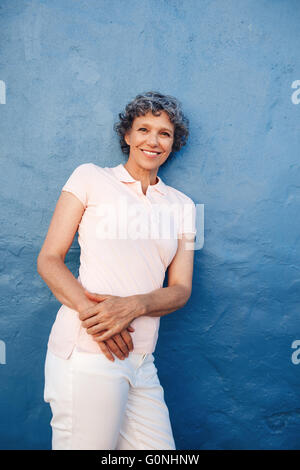 Porträt des entspannten Reife Dame zu einer blauen Wand gelehnt. Glücklich mittleren gealterten weiblichen stehen vor blauem Hintergrund Stockfoto