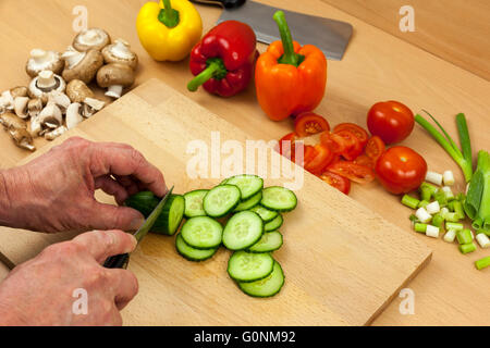 Nahaufnahme Schuss eine Köche Hände schneiden eine englische Gurke auf ein Schneidbrett aus Holz zur Seite eine Auswahl an geschnittenen Mischgemüse Stockfoto