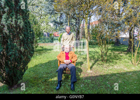 Vater und Sohn, älterer Mann sitzt auf einem Stuhl und Mann mittleren Alters zusammen im Garten Stockfoto