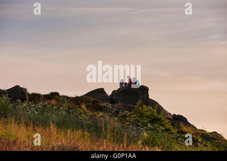 Familie von vier sitzen oben, hoch auf einem Felsvorsprung, um den Blick bei Sonnenuntergang - Otley Chevin, North Yorkshire, England. Stockfoto