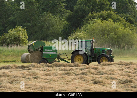 Great Ouseburn, North Yorkshire, GB, UK-runden Ballen ablegen und Fallen von einer Ballenpresse mit einem grünen farm Traktor in einem Feld gezogen, die silage. Stockfoto