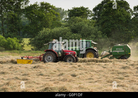 Zwei landwirtschaftliche Traktoren Silageherstellung in einem Feld bei großen Ouseburn, North Yorkshire, England - eine grüne ziehen eine Ballenpresse, einem roten ziehen eine Harke. Stockfoto