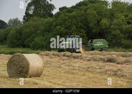 Heu oder Silage, Great Ouseburn, North Yorkshire, England - Rundballen in Feld mit grünen Traktor fahren & Arbeiten über, Ziehen einer Ballenpresse. Stockfoto