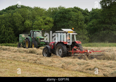 Zwei landwirtschaftliche Traktoren Silageherstellung in einem Feld bei großen Ouseburn, North Yorkshire, England - die grünen eine ziehen eine Ballenpresse, die rote Linie schleppen einen Rechen. Stockfoto