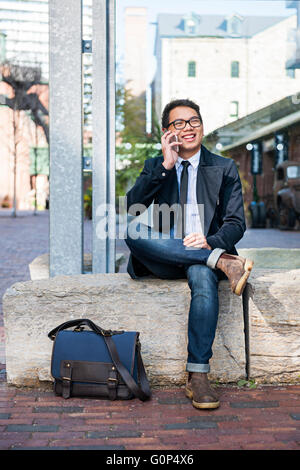 Glückliche junge asiatische Geschäftsmann telefonieren mit Handy draußen sitzen auf Stadt Straße Stockfoto