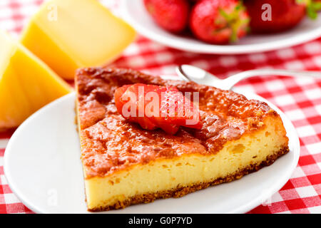 Nahaufnahme einer Platte mit einem Stück hausgemachter Käsekuchen garniert mit in Scheiben geschnittenen Erdbeeren auf einem Tisch mit einer karierten Tischdecke Stockfoto