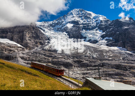 Zug der Jungfraubahn läuft in Richtung Jungfraujoch unter dem Monch und Eiger Gletscher in der Nähe der Station Eigergletscher auf Berner Stockfoto