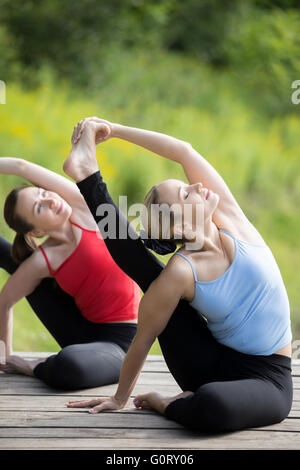 Yogastunde: zwei ruhige junge schöne Frauen arbeiten auf Sommertag draußen im Park, im Parivrtta Surya Yantrasana sitzen Stockfoto