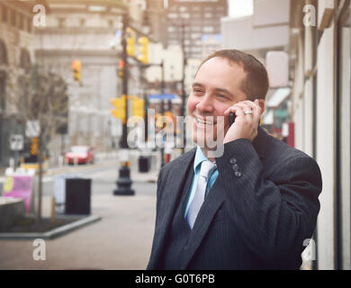 Ein Business-Mann ist auf einem Handy außerhalb in einer Stadt trägt einen Anzug und Lächeln für einen Erfolg oder Kommunikation Idee sprechen. Stockfoto