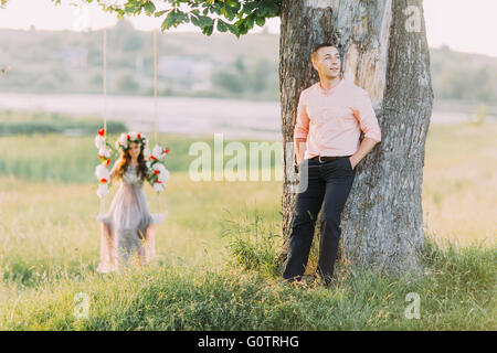 Hübsche junge Blondine trägt lila Kleid und Kranz in Gange während ihrer hübschen Freund Stand in der Nähe Baum sitzen Stockfoto
