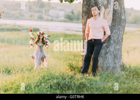 Hübsche junge Blondine trägt lila Kleid und Kranz in Gange während ihrer hübschen Freund Stand in der Nähe Baum sitzen Stockfoto