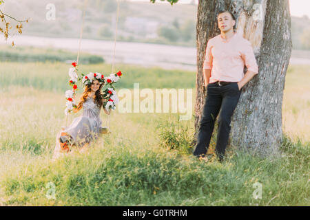 Hübsche junge Blondine trägt lila Kleid und Kranz in Gange während ihrer hübschen Freund Stand in der Nähe Baum sitzen Stockfoto