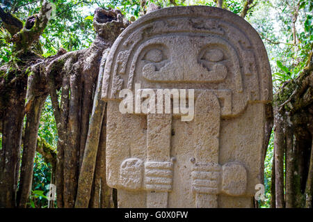 Eine geheimnisvolle Statue einer männlichen Person steht im Regenwald neben einem alten Baum mit großen Wurzeln. Die Statuen von San Agustin Stockfoto