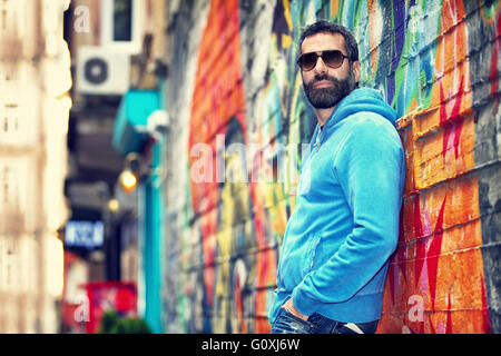 Gut aussehender Mann mit stilvollen Sonnenbrille, Stand in der Nähe schöne bunte Wand auf der Straße, urban Fashion-Look, Leben in der Stadt Stockfoto