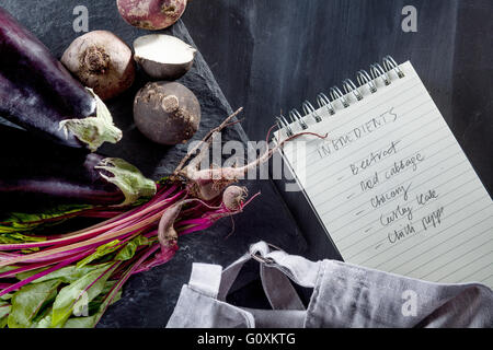 Rüben, Auberginen, schwarze Rüben und Rübenblätter mit Schürze und Notebook auf der Seite Ansicht von oben Stockfoto