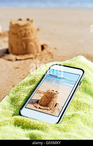 eine Smartphone mit einem Bild von mir eine Sandburg in seinem Bildschirm gedreht platziert auf einem grünen Handtuch auf dem Sand des Strandes und der Stockfoto