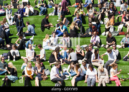 Eine große Gruppe von Freunden, Kollegen und Kolleginnen und Kollegen genießen Sie ihr Mittagessen brechen in der Sonne auf einer grasbewachsenen Bank in der Stadt. Stockfoto